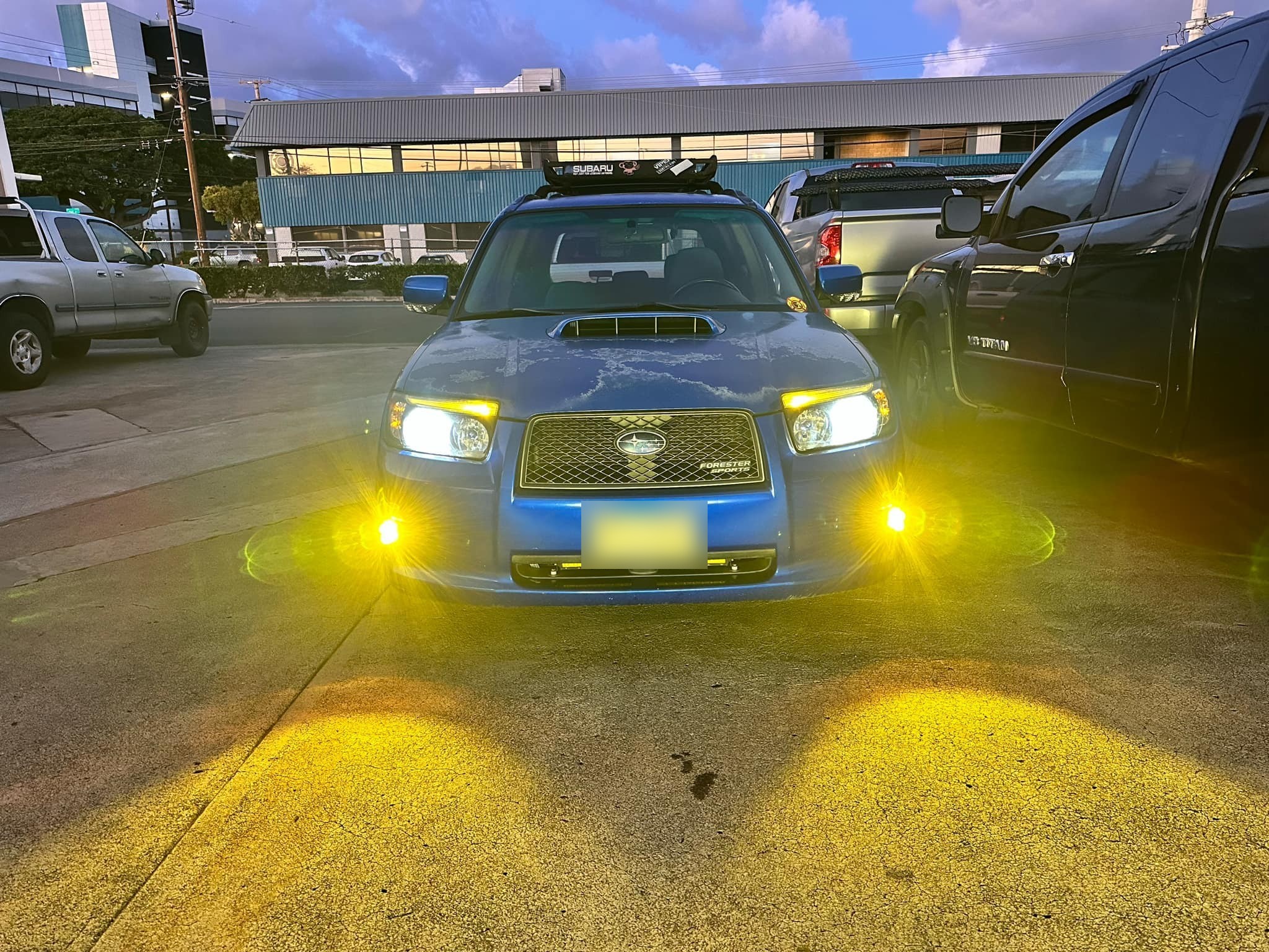 The blue Forester at dusk with both amber fog lights throwing beam patterns onto the pavement